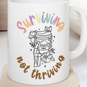 May include: White ceramic mug with the words "Surviving not thriving" in a colorful, whimsical font. The mug features a black and white illustration of a coffee cup with a skeleton design. The mug sits on a beige coaster.