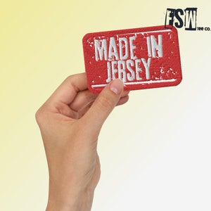 Może przedstawiać: Czerwona łatka z tkaniny z białym haftem, na której widnieje napis "Made in Jersey".