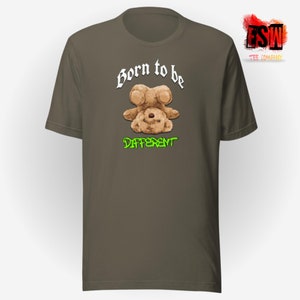 Op de afbeelding: Een bruin t-shirt met een witte afbeelding van een ondersteboven teddybeer met de tekst "Born to be Different" in wit en groen.