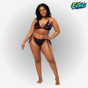 Puede incluir: Un conjunto de bikini negro con un estampado abstracto rosa. El top triangular y la braguita tienen lazos. La modelo sonríe frente a un fondo blanco. El logo "ESW Tee Co" está en la esquina superior derecha.