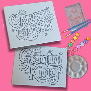 Könnte beinhalten: Zwei leere weiße Leinwände mit schwarzen Umrissen der Wörter "Cancer Queen" und "Gemini King" mit einer Krone über jedem Wort. Die Leinwände sind von Malutensilien umgeben, darunter eine Farbpalette, Pinsel und kleine Farbflaschen.
