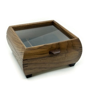 May include: A dark brown wooden box with a glass top and a small wooden handle. The box has a wood grain pattern and is sitting on a white surface.