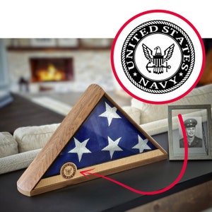 May include: A wooden flag display case with a folded American flag inside. The case has a glass front and a brass plaque with the United States Navy seal on the bottom.
