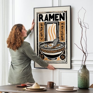 Pode incluir: Uma mulher pendura um p&ocirc;ster emoldurado em preto com uma ilustra&ccedil;&atilde;o vintage de uma tigela de macarr&atilde;o ramen. O p&ocirc;ster tem a palavra "Ramen" em letras grandes no topo e as palavras "Safety Matches" do lado direito. O p&ocirc;ster est&aacute; em uma parede branca.