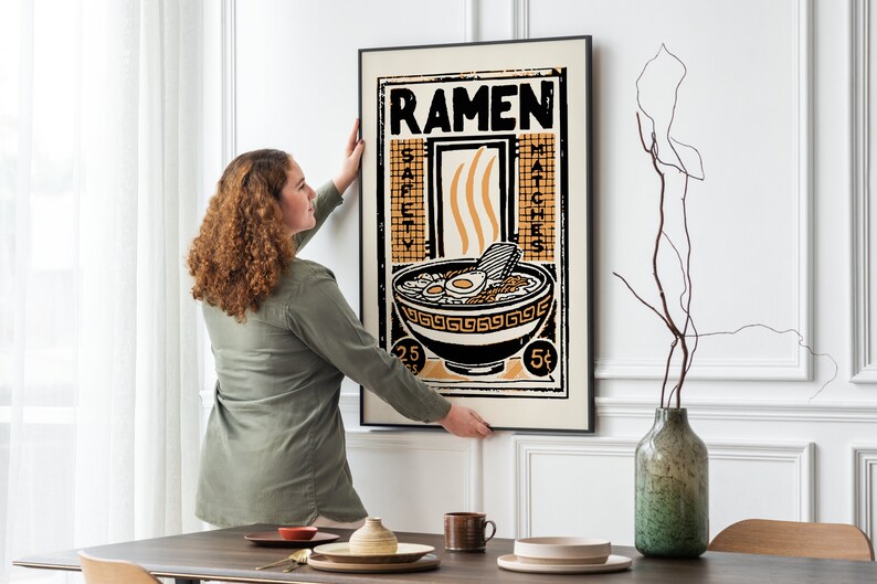 Pode incluir: Uma mulher pendura um p&ocirc;ster emoldurado em preto com uma ilustra&ccedil;&atilde;o vintage de uma tigela de macarr&atilde;o ramen. O p&ocirc;ster tem a palavra "Ramen" em letras grandes no topo e as palavras "Safety Matches" do lado direito. O p&ocirc;ster est&aacute; em uma parede branca.