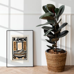 Pode incluir: Um p&ocirc;ster emoldurado em preto com uma ilustra&ccedil;&atilde;o vintage de uma tigela de macarr&atilde;o ramen. O p&ocirc;ster tem a palavra "RAMEN" em letras pretas em negrito no topo e as palavras "SAFETY MATCHES" &agrave; esquerda e &agrave; direita da tigela. O p&ocirc;ster est&aacute; pendurado em uma parede branca ao lado de uma planta de borracha em vaso.