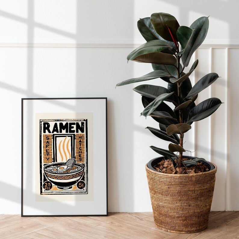 Pode incluir: Um p&ocirc;ster emoldurado em preto com uma ilustra&ccedil;&atilde;o vintage de uma tigela de macarr&atilde;o ramen. O p&ocirc;ster tem a palavra "RAMEN" em letras pretas em negrito no topo e as palavras "SAFETY MATCHES" &agrave; esquerda e &agrave; direita da tigela. O p&ocirc;ster est&aacute; pendurado em uma parede branca ao lado de uma planta de borracha em vaso.