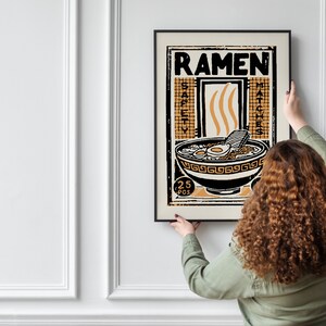 Pode incluir: Uma pessoa est&aacute; a pendurar um poster emoldurado numa parede branca. O poster &eacute; preto e branco com uma ilustra&ccedil;&atilde;o amarela e laranja de uma tigela de ramen. O poster diz "Ramen" e "25 pcs" num estilo vintage.