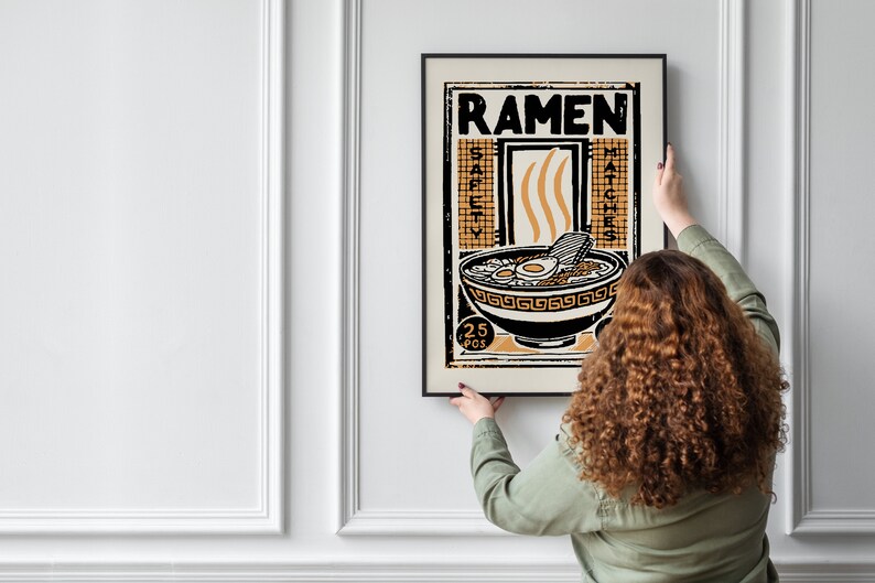 Pode incluir: Uma pessoa est&aacute; a pendurar um poster emoldurado numa parede branca. O poster &eacute; preto e branco com uma ilustra&ccedil;&atilde;o amarela e laranja de uma tigela de ramen. O poster diz "Ramen" e "25 pcs" num estilo vintage.