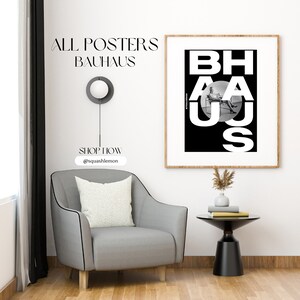May include: A black and white poster with the text "BAHAUS" in bold white letters. The letters are arranged in a circle with a person sitting in a chair in the center of the circle. The poster is framed in a light wood frame and is hanging on a white wall.