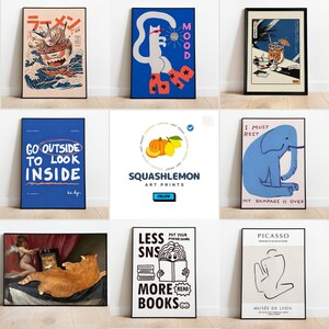 May include: A collage of nine different art prints featuring various designs, including a cat in sunglasses, a bowl of ramen, a blue elephant, and a cat looking at a painting. The prints are all framed in black frames and are displayed on a white wall.