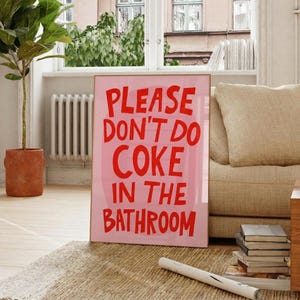 Op de afbeelding: Een ingelijste roze poster met de tekst "PLEASE DON'T DO COKE IN THE BATHROOM" in rode, vette letters. De poster leunt tegen een beige bank en een raam met uitzicht op een gebouw.