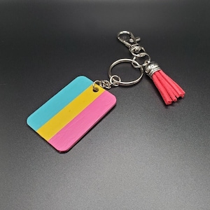 Op de afbeelding: Een sleutelhanger met een blauw, geel en roze rechthoekig label bevestigd aan een zilveren sleutelring en een rode kwast.