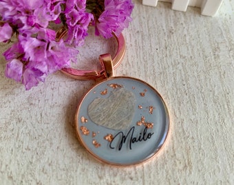 May include: A rose gold keychain with a round, gray resin charm. The charm features a heart-shaped piece of pressed foliage and gold glitter. The word "Maile" is written in black cursive on the charm.