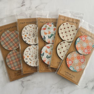 May include: Six sets of car coasters with various patterns. Each set includes two coasters. The patterns include plaid, floral, butterflies, hearts, and geometric designs. The coasters are made of wood and have a cork backing.