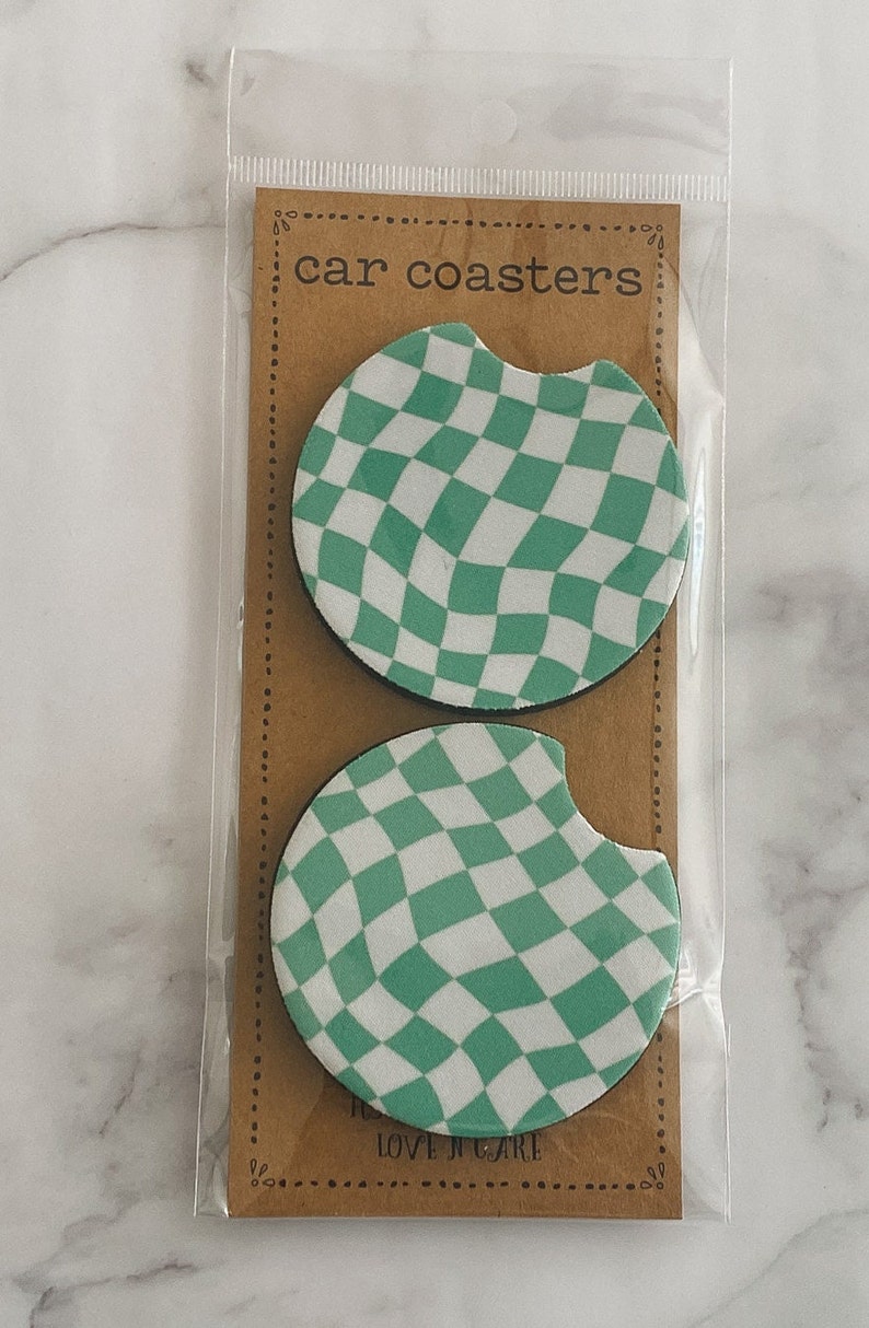 Pu&ograve; includere: Due sottobicchieri per auto a scacchi verdi e bianchi in una confezione di plastica. La confezione ha uno sfondo marrone con il testo "car coasters" stampato su di essa.