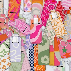 May include: A collection of colorful keychains with various patterns, including abstract swirls, flowers, stars, and geometric designs. The keychains are made of fabric and have silver metal rings.