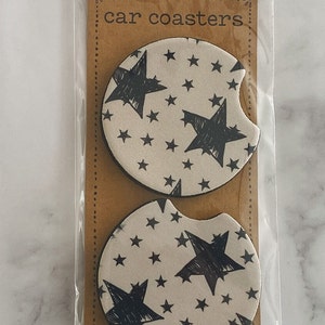 Pu&ograve; includere: Due sottobicchieri rotondi per auto con sfondo bianco e un motivo a stelle nere. I sottobicchieri sono confezionati in un sacchetto di plastica trasparente con un retro in cartone marrone che dice "car coasters" e "love n care".