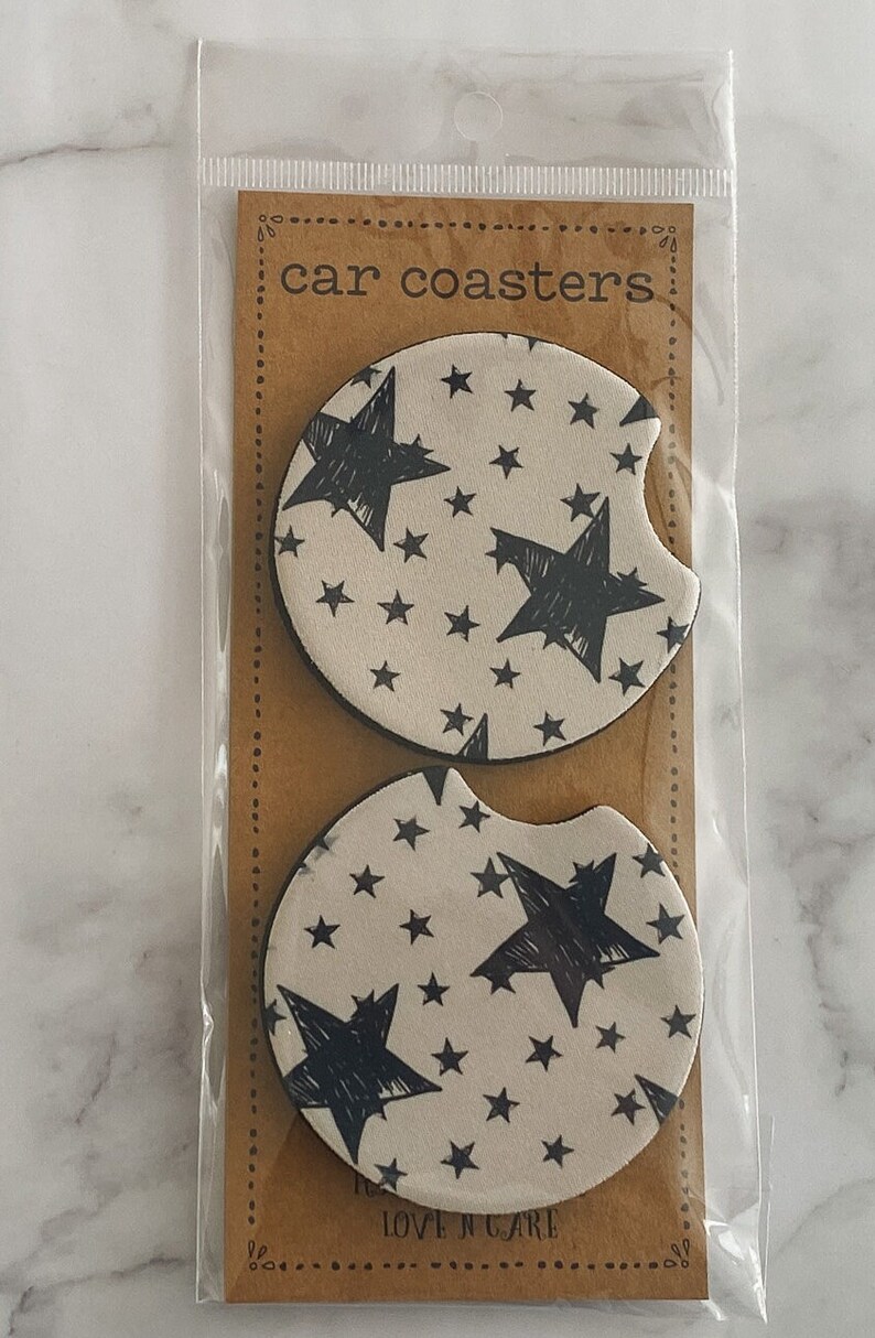 Pu&ograve; includere: Due sottobicchieri rotondi per auto con sfondo bianco e un motivo a stelle nere. I sottobicchieri sono confezionati in un sacchetto di plastica trasparente con un retro in cartone marrone che dice "car coasters" e "love n care".