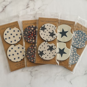 May include: Set of six car coasters featuring various patterns. The coasters are made of cork and have a round shape. The patterns include stars, floral designs, and a blue and white geometric pattern. The coasters are packaged in clear plastic bags and labeled "car coasters".