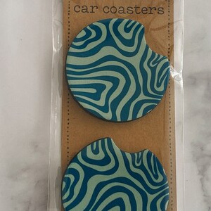 Pu&ograve; includere: Due sottobicchieri per auto con un motivo a spirale blu e verde in una busta di plastica trasparente con un cartoncino marrone su cui &egrave; scritto "car coasters".