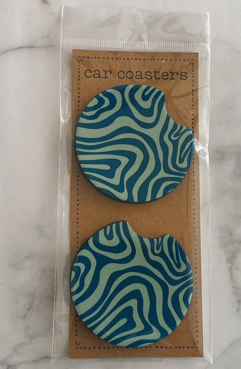 Pu&ograve; includere: Due sottobicchieri per auto con un motivo a spirale blu e verde in una busta di plastica trasparente con un cartoncino marrone su cui &egrave; scritto "car coasters".