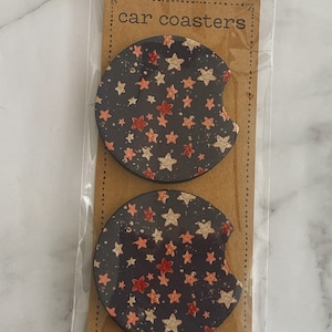 Pu&ograve; includere: Due sottobicchieri rotondi per auto con sfondo nero e un motivo a stelle bianche e rosa. I sottobicchieri sono confezionati in un sacchetto di plastica trasparente con un'etichetta di carta marrone che dice "car coasters".