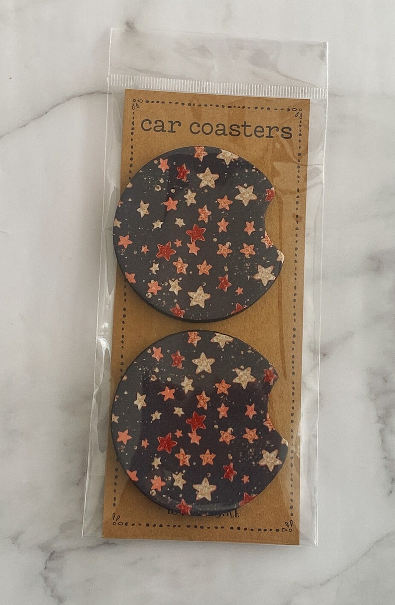 Pu&ograve; includere: Due sottobicchieri rotondi per auto con sfondo nero e un motivo a stelle bianche e rosa. I sottobicchieri sono confezionati in un sacchetto di plastica trasparente con un'etichetta di carta marrone che dice "car coasters".