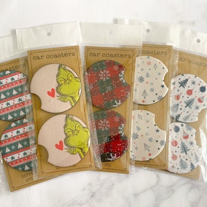 May include: Three sets of car coasters featuring a Christmas theme. One set has a red and green plaid design, another has a white background with blue and red ornaments, and the last set has a Grinch design with a pink background.