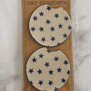 Pu&ograve; includere: Due sottobicchieri rotondi per auto con sfondo bianco e un motivo a stelle blu. I sottobicchieri sono confezionati in un sacchetto di plastica trasparente con un retro in cartone marrone che dice "car coasters" e "handmade with love n care".