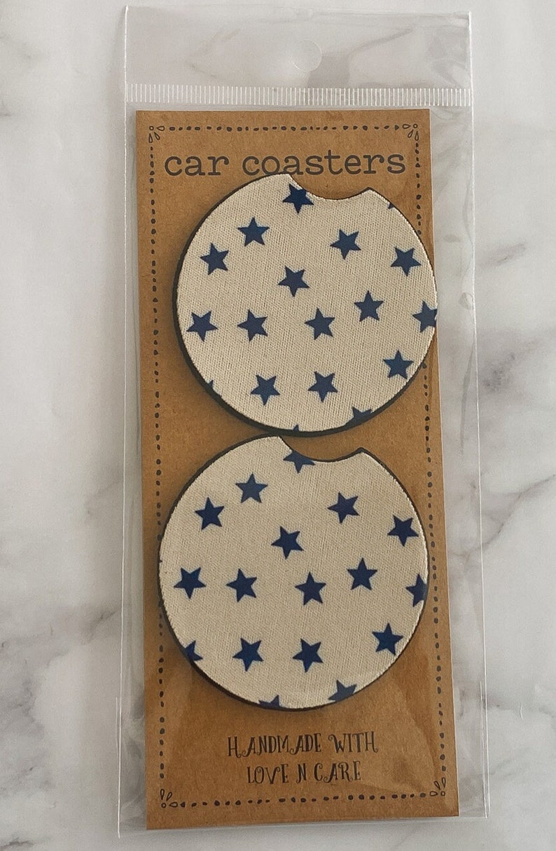 Pu&ograve; includere: Due sottobicchieri rotondi per auto con sfondo bianco e un motivo a stelle blu. I sottobicchieri sono confezionati in un sacchetto di plastica trasparente con un retro in cartone marrone che dice "car coasters" e "handmade with love n care".