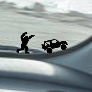 May include: A black silhouette of a Bigfoot running towards a black silhouette of a jeep. The image is on a car window.