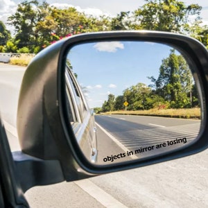 May include: A rearview mirror with the text "objects in mirror are losing" printed on the bottom. The mirror is reflecting a road with trees and a car in the distance.