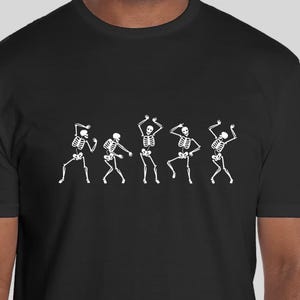 May include: Black t-shirt with a white graphic of five skeletons dancing. The skeletons are in a line and are all facing the same direction. They are all wearing different poses.