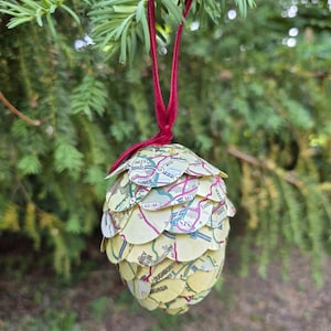 May include: A handmade Christmas ornament shaped like a pinecone, crafted from map paper. The ornament is suspended by a red velvet ribbon and hangs against a backdrop of green foliage. The map paper has shades of yellow, green, and pink.