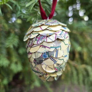May include: A handmade Christmas ornament shaped like a pinecone. The ornament is constructed from layered, scalloped pieces of vintage map paper. A red ribbon is attached for hanging. The ornament is displayed against a backdrop of green pine needles.