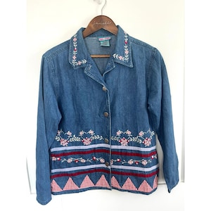 May include: A blue denim jacket featuring a collar and front button closure. The garment is decorated with embroidered pink floral patterns on the collar and mid-section, horizontal red and light blue stripes, and a red and white gingham hem.