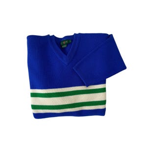 Puede incluir: Un suéter azul con cuello en V con rayas blancas y verdes en la parte inferior. El suéter tiene mangas largas y una etiqueta que dice "J.Crew". El suéter está doblado y se muestra sobre un fondo blanco.