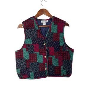 Op de afbeelding: Een patchwork vest in verschillende patronen en kleuren, waaronder rood geruit, groen bloemenpatroon en marineblauw. Het vest heeft een V-hals, goudkleurige knopen en een gewatteerde voering. Het vest hangt aan een houten hanger.