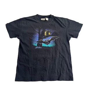 May include: A black short-sleeve t-shirt featuring a graphic print of a bat flying in front of a tree with a lit-up treehouse at night. The L.L.Bean logo is printed in blue below the bat. The shirt has a crew neckline and a brand label at the collar.