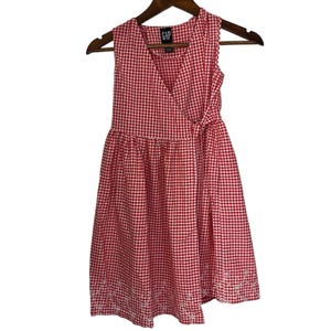 May include: A sleeveless red and white gingham dress with a wrap-style bodice and flared skirt. The dress has a tie closure at the waist and white embroidery on the hem. The GAP brand label is visible. The dress hangs on a wooden hanger. The dress is suitable for a child.