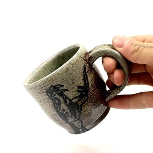 Puede incluir: Una taza de cerámica con esmalte moteado en tonos verde claro y marrón, que presenta un gráfico oscuro y abstracto dibujado a mano en el lateral. La taza tiene un asa gruesa y curva con un acabado terroso y texturizado.