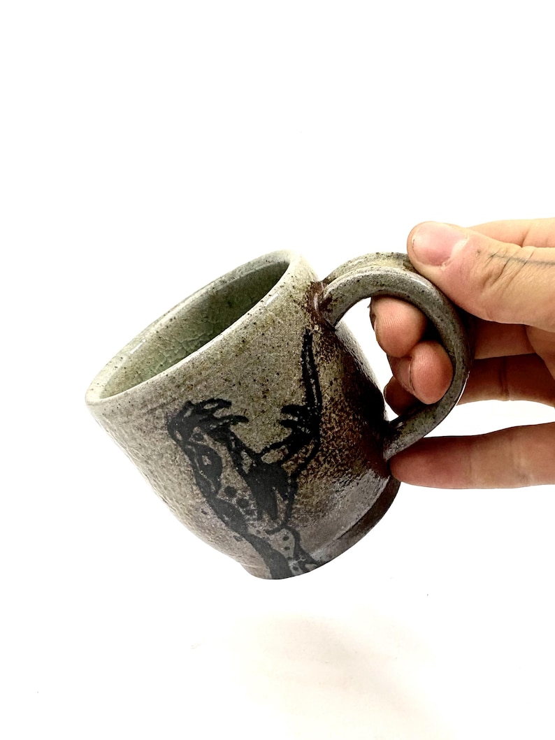 May include: A ceramic mug with a speckled light green and brown glaze, featuring a dark, abstract hand-drawn graphic on the side. The mug has a thick, curved handle and a textured, earthy finish with visible crackle patterns in the glaze.