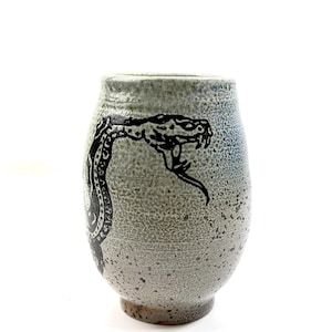 May include: A ceramic tumbler with a speckled light grey and blue glaze featuring a black graphic illustration of a coiled snake with its mouth open, showing fangs and a forked tongue. The vessel has a rounded body and a narrow base.