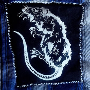 May include: A white rat with a long tail is depicted on a black fabric patch. The rat is facing the right side of the image and has a menacing expression. The patch is sewn onto a blue denim fabric.