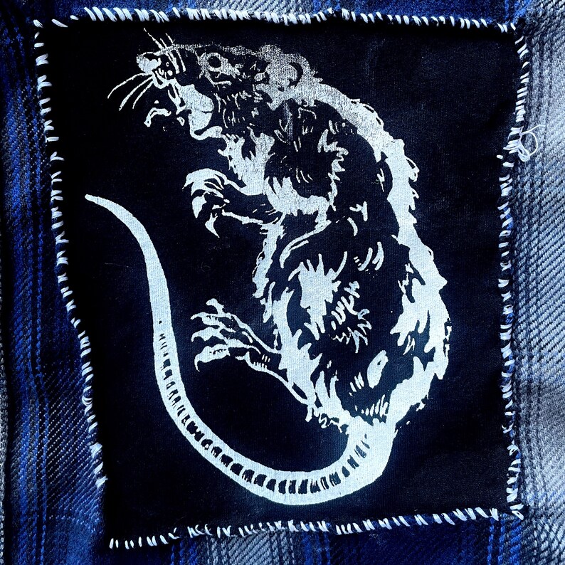 May include: A white rat with a long tail is depicted on a black fabric patch. The rat is facing the right side of the image and has a menacing expression. The patch is sewn onto a blue denim fabric.