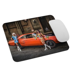 May include: Two women in vintage clothing stand next to a bright orange 1970s muscle car. The car has a black stripe down the side and the words "Six Pack" on the back. The women are wearing white go-go boots and the car is parked in front of a brick building.