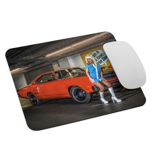 May include: A bright orange 1969 Dodge Charger with a woman in a blue and white dress standing next to it in a parking garage. The car has black racing stripes and black wheels with red trim. The woman is wearing white boots and is looking at the camera.