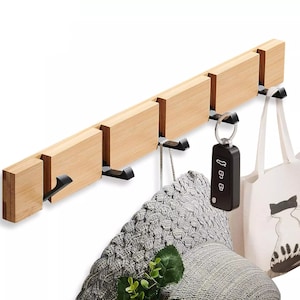 May include: A wooden wall-mounted coat rack with five hooks. The rack is made of light brown wood and has a natural finish. A hat, a tote bag, and a set of keys are hanging on the hooks.