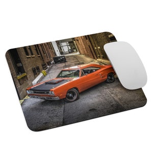 May include: Mousepad featuring an orange classic car with a black hood, parked in a narrow alley. The image includes a white computer mouse. The car is the focal point, set against a backdrop of brick buildings.